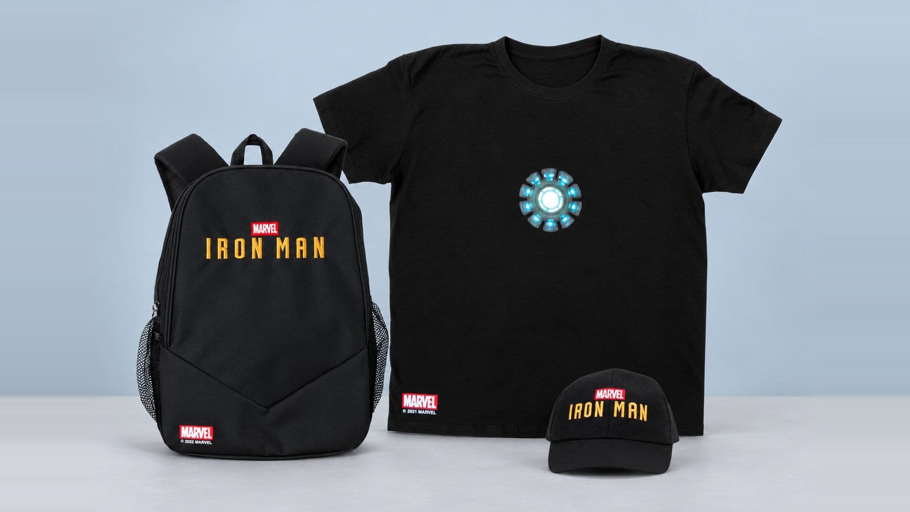 Licensed character merchandise on a blue-grey studio backdrop: a black backpack, a black T-shirt with a glowing circular chest print and a black baseball cap